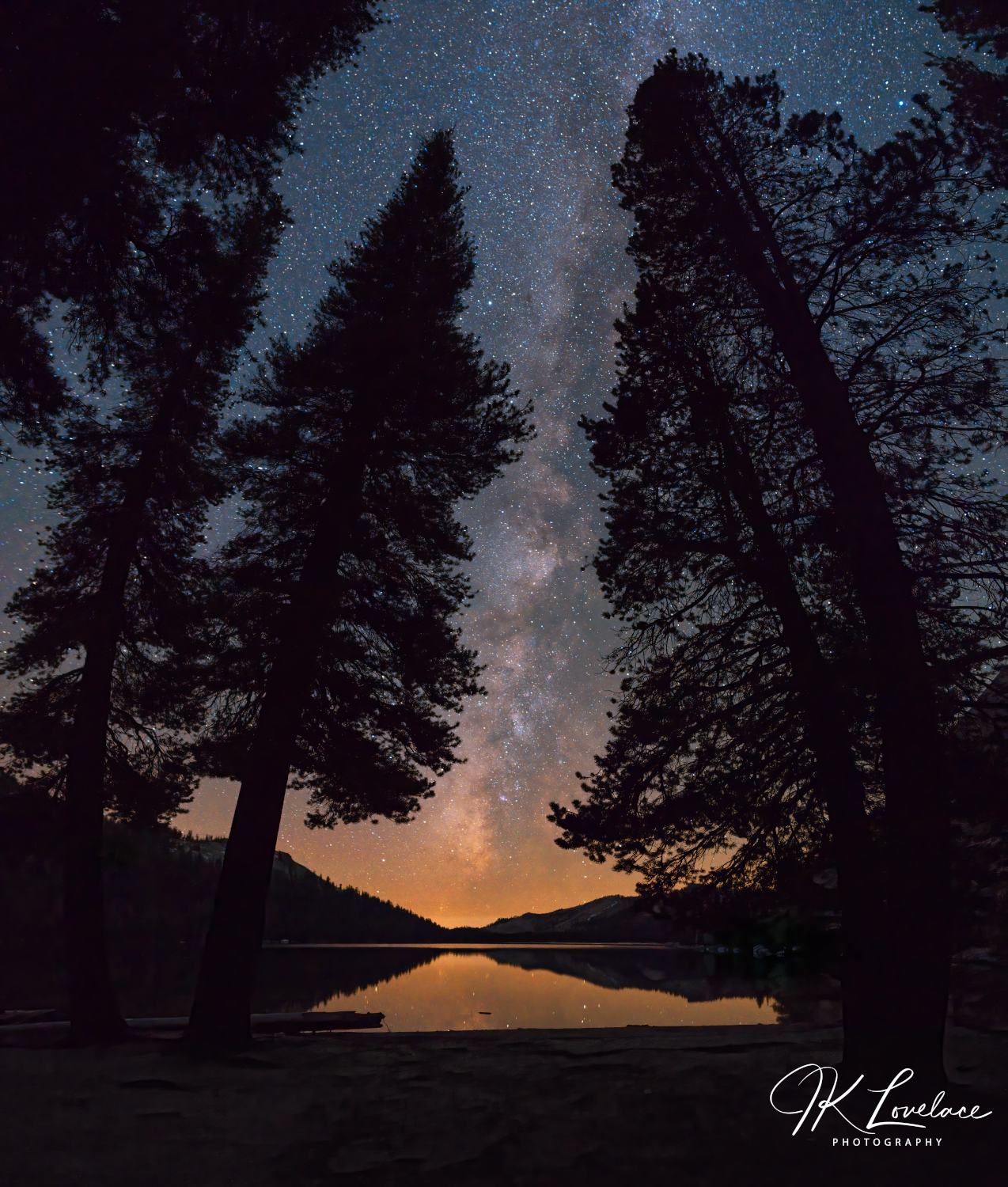 A png of the nightscape photograph titled "All is Calm" shot by landscape photographer J K Lovelace featuring a view of Tenaya Lake at night with tree silhouettes and the Milky Way Galaxy.