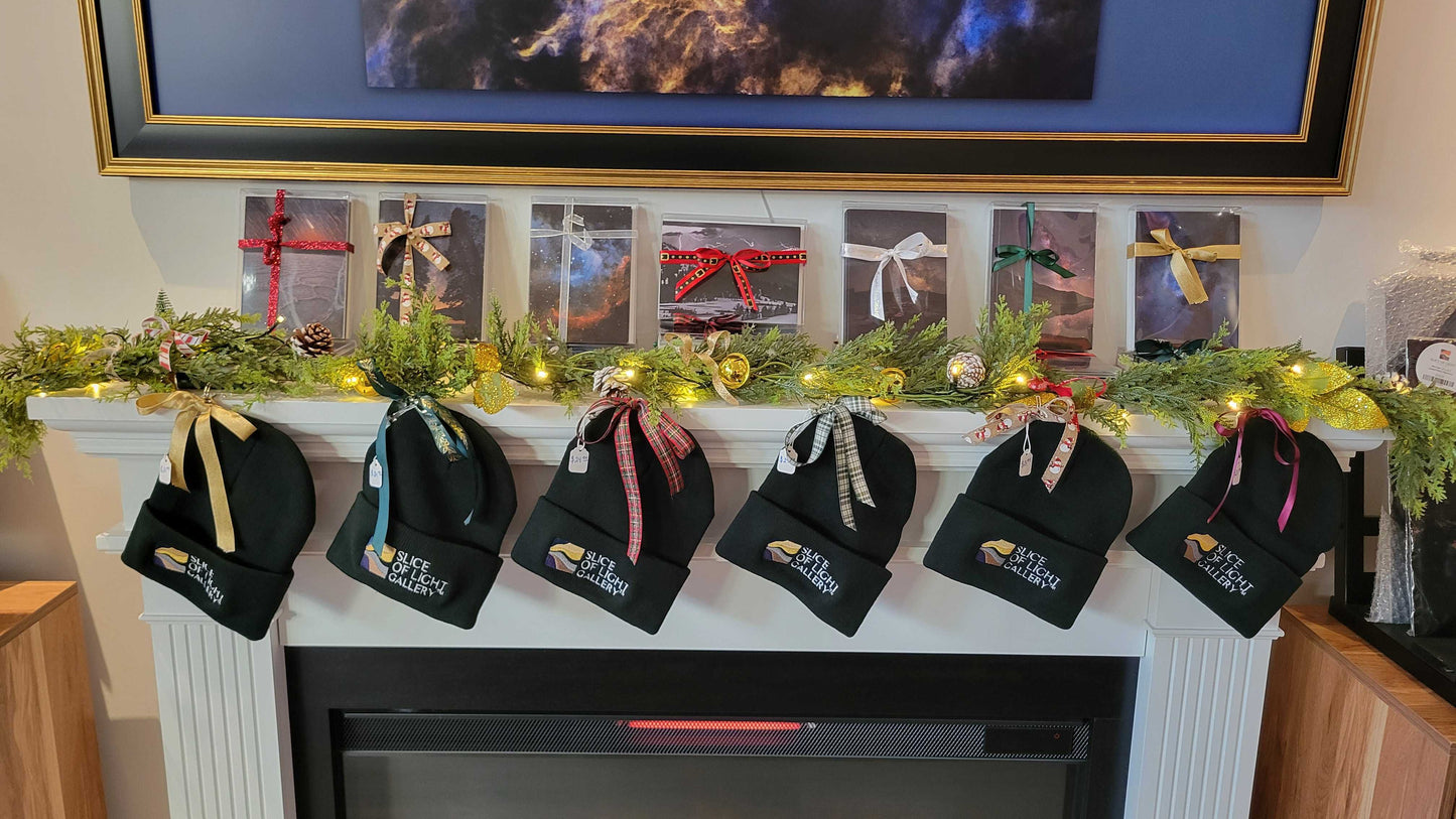 A photograph showing a bunch of black beanies and greeting card sets lined up on a fireplace mantel showing they are for sale at Slice of Light Gallery.