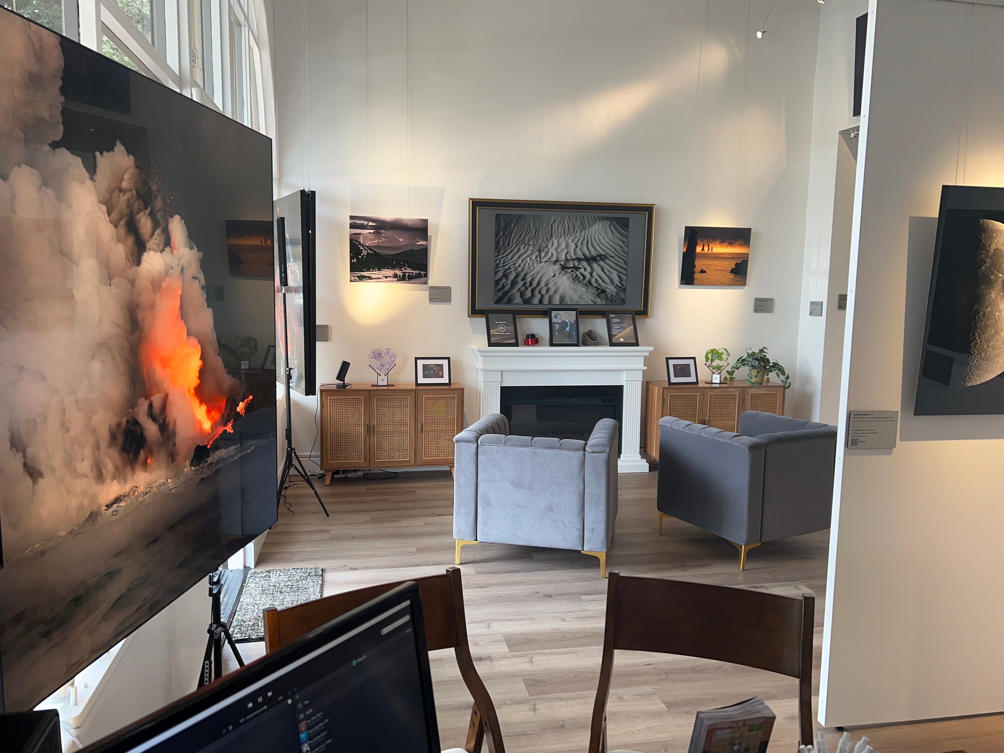 A photograph of the interior view of the gallery taken from the vantage point of the sales desk, showing multiple metal prints, tv, and the fireplace.