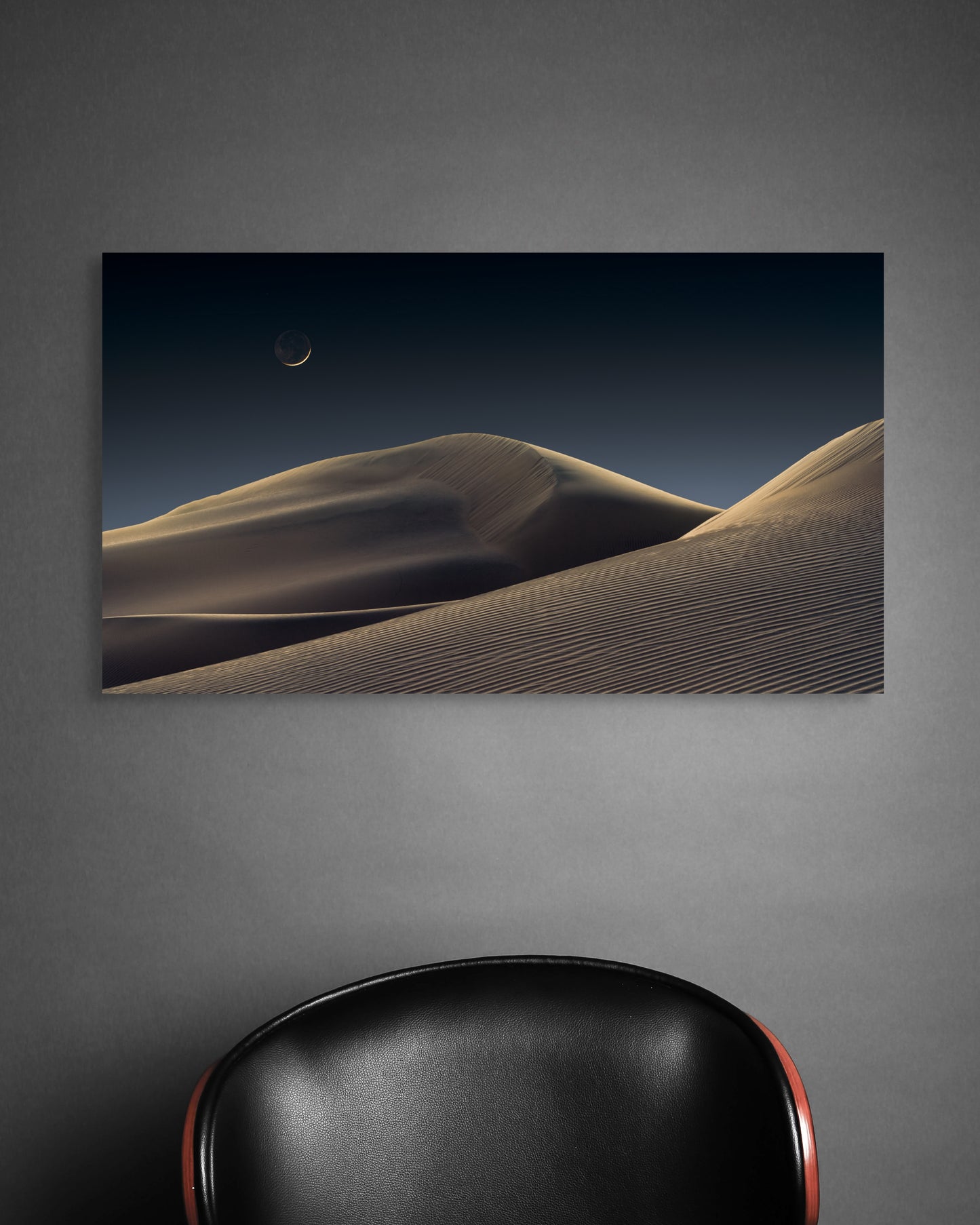 A wall art mockup featuring the photograph titled "Luna Dunes" by landscape photographer J K Lovelace as a 30 inch wide metal print with inset frame on display on dark wall above black leather chair.