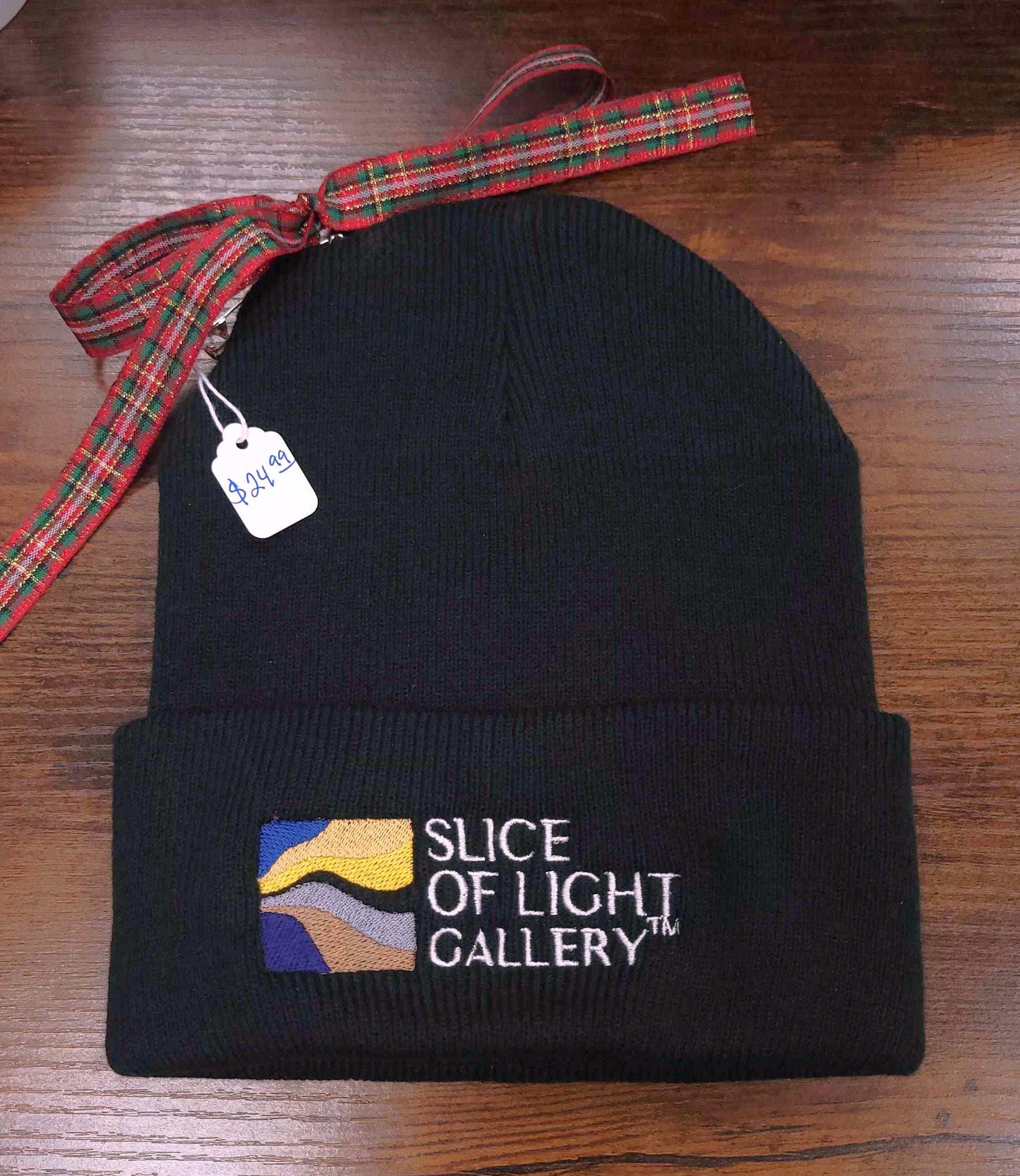 A photograph of a black beanie embroidered with the Slice of Light Gallery logo paired with a price tag and xmas ribbon