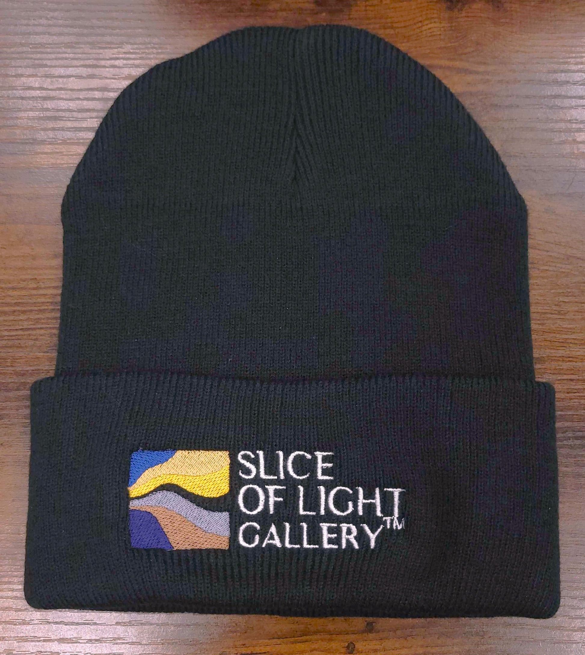 A photograph of a black beanie embroidered with the Slice of Light Gallery logo laying flat on a desk