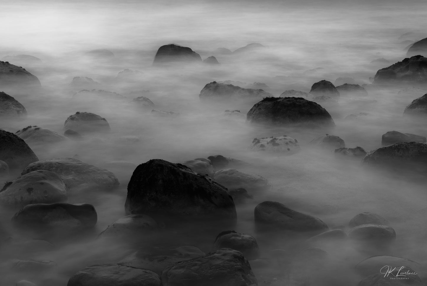A jpg of a black and white seascape photograph titled "Neoteric Soup" shot by landscape photographer J K Lovelace featuring Santa Barbara Coast round boulders and mist.