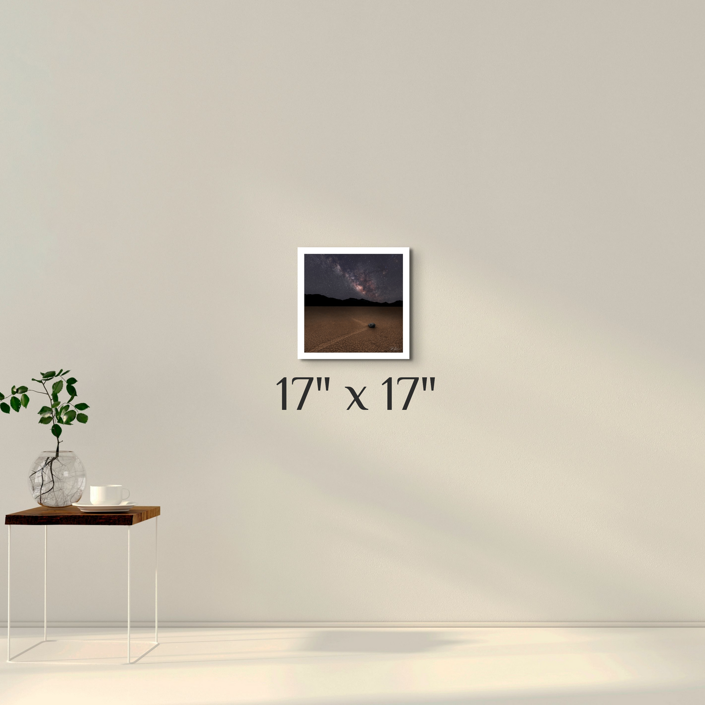 A framed artwork featuring a nightscape image of a dry lakebed with a milky way, displayed on a wall above a minimalist table with a plant and a vase.