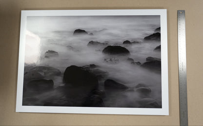 Framed black and white photograph of rocks and water on a beige wall with a ruler for scale.