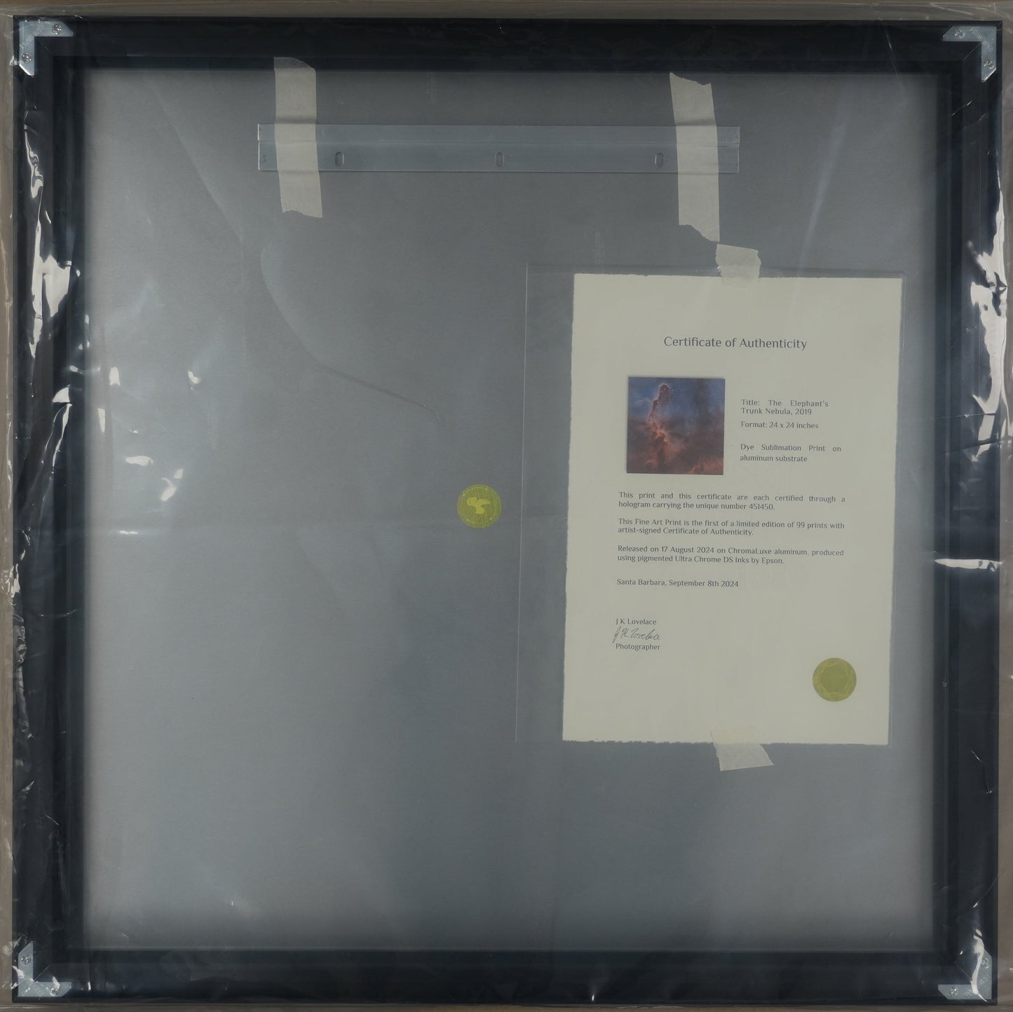 Framed artwork with a certificate inside, wrapped in plastic on a cardboard surface.