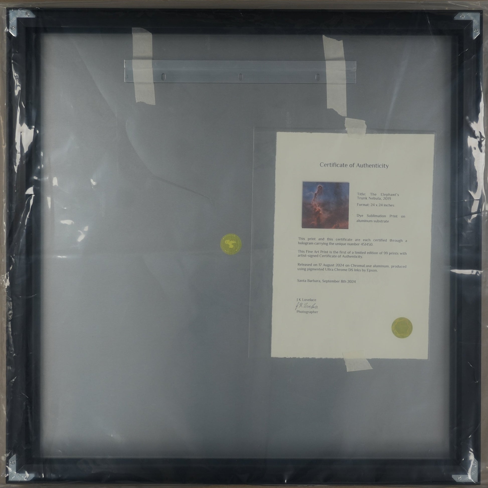 Framed artwork with a certificate inside, wrapped in plastic on a cardboard surface.