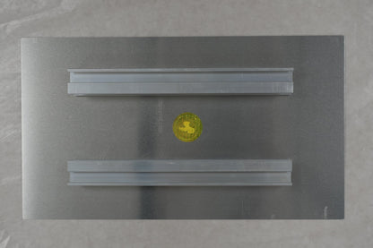 Rectangular metallic object with a yellow circle on a textured surface, with a ruler at the bottom.