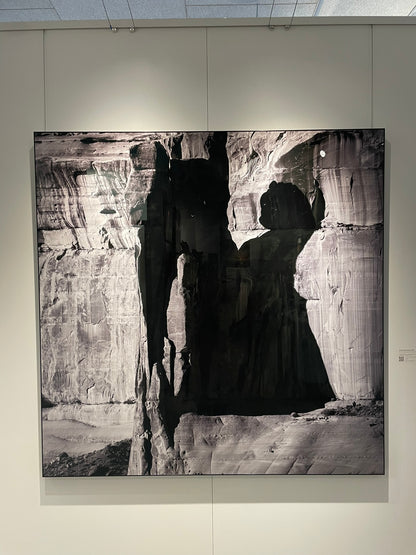 Black and white photograph of a person in a dark coat standing in front of a rocky landscape.