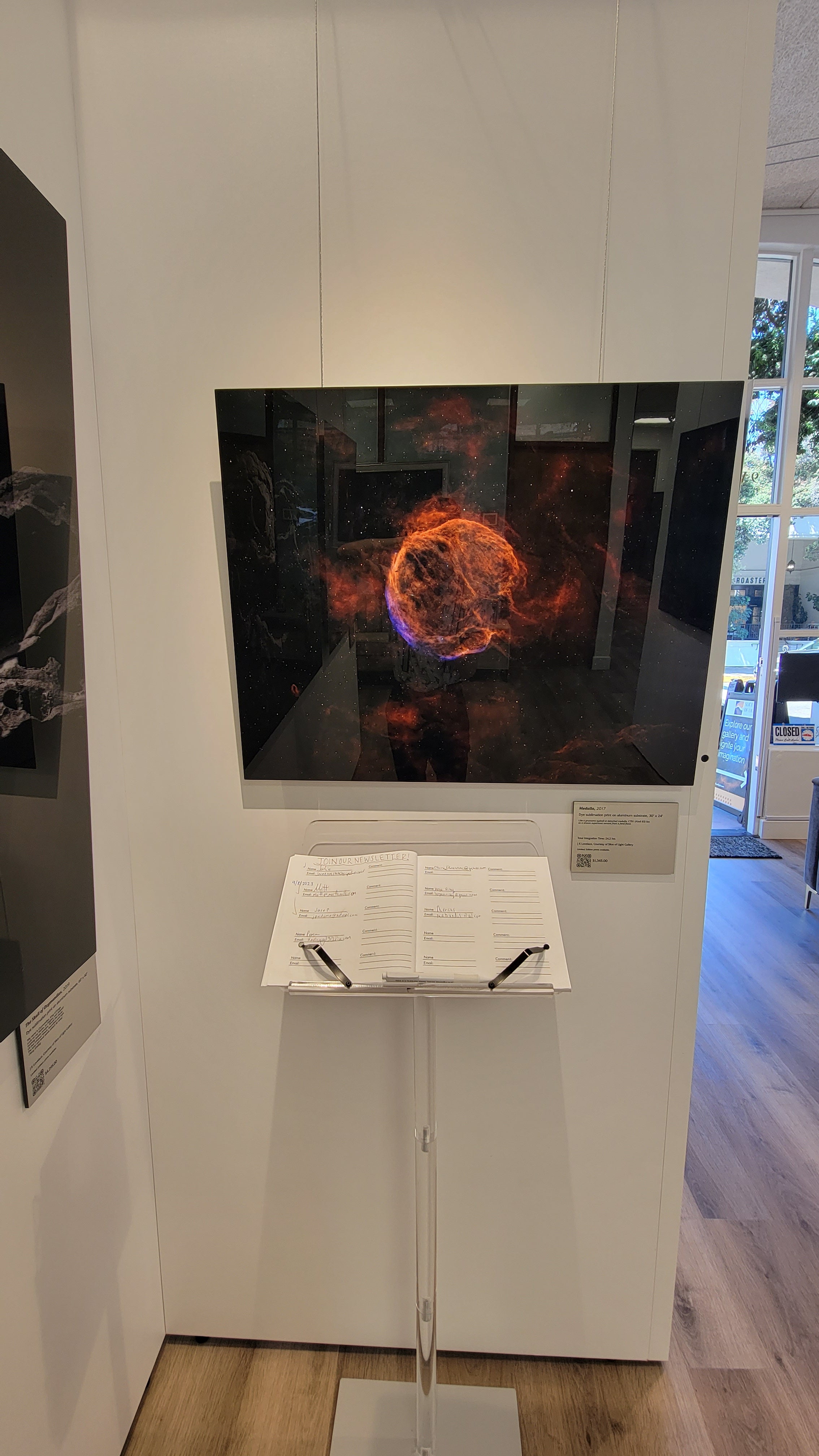 A photograph showing the metal print titled "Medulla" on display on a wall partition above a sign in list on a pedestal within Slice of Light Gallery.