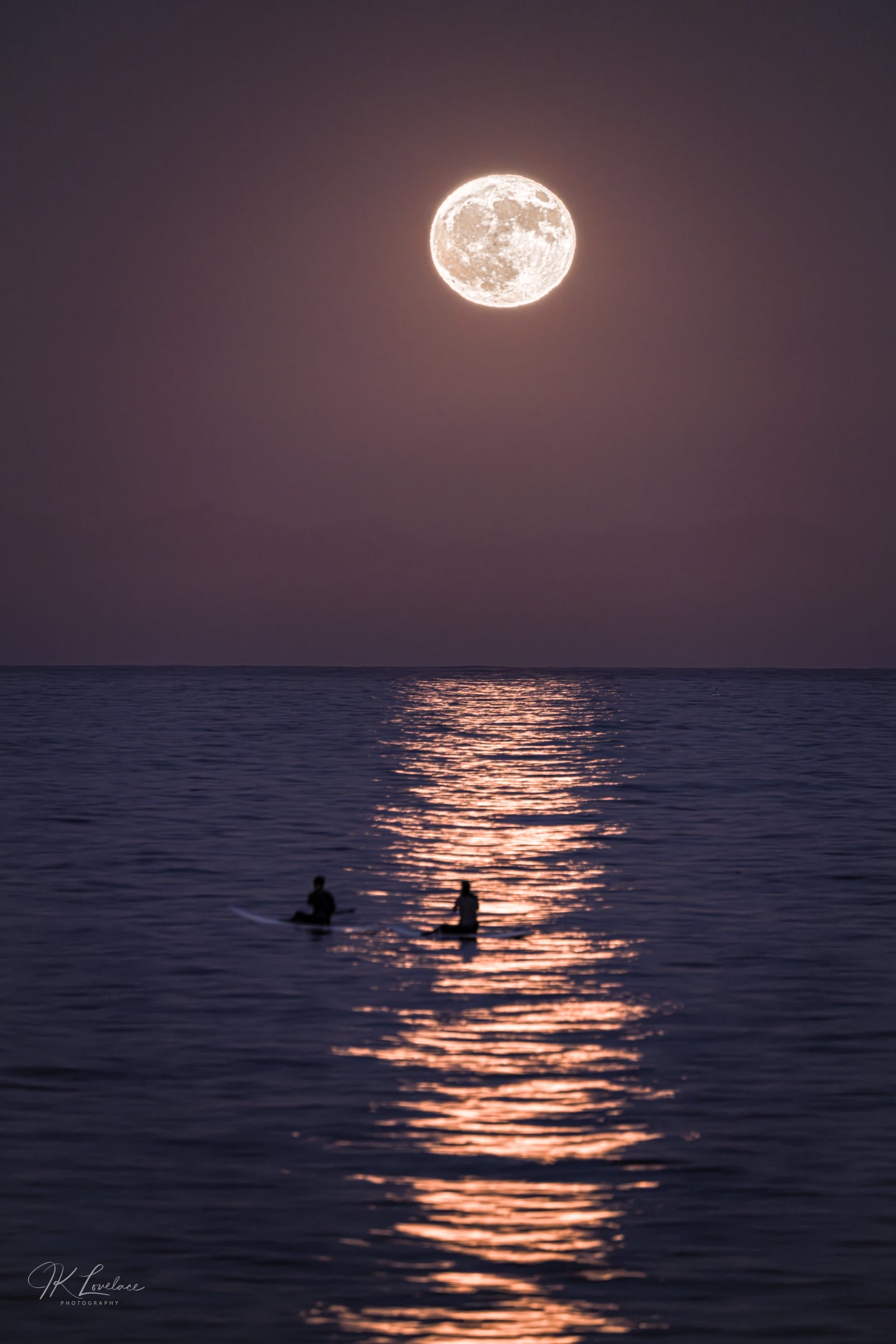 A jpg of the photograph titled "Moon Watching" shot by landscape photographer J K Lovelace, featuring a night photograph of surfboarders in the ocean and a full moon.