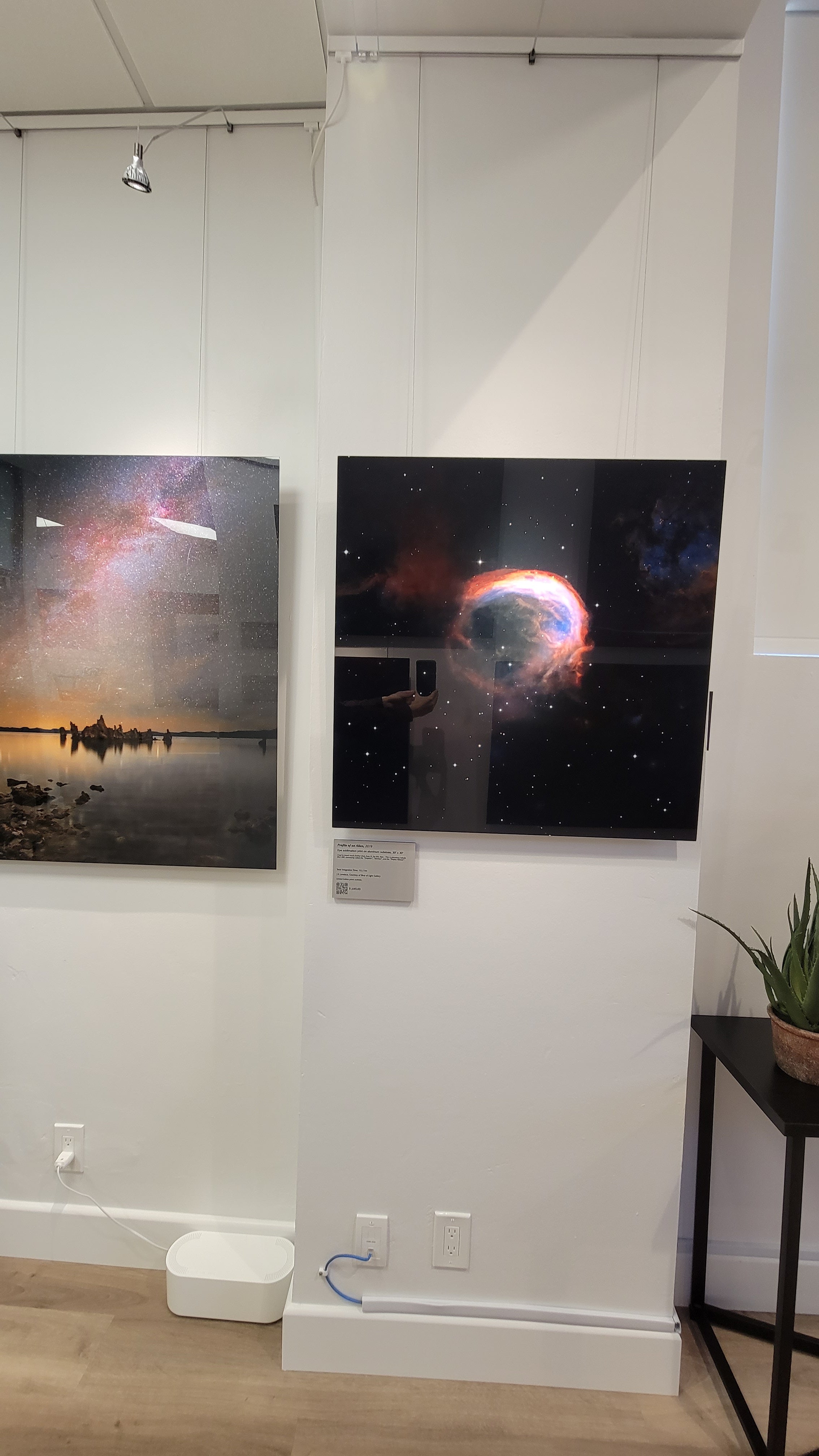 A photograph of the gallery interior showing the metal prints titled "Profile of an Alien" and "Mono Midnight" on display in the cosmic cove.