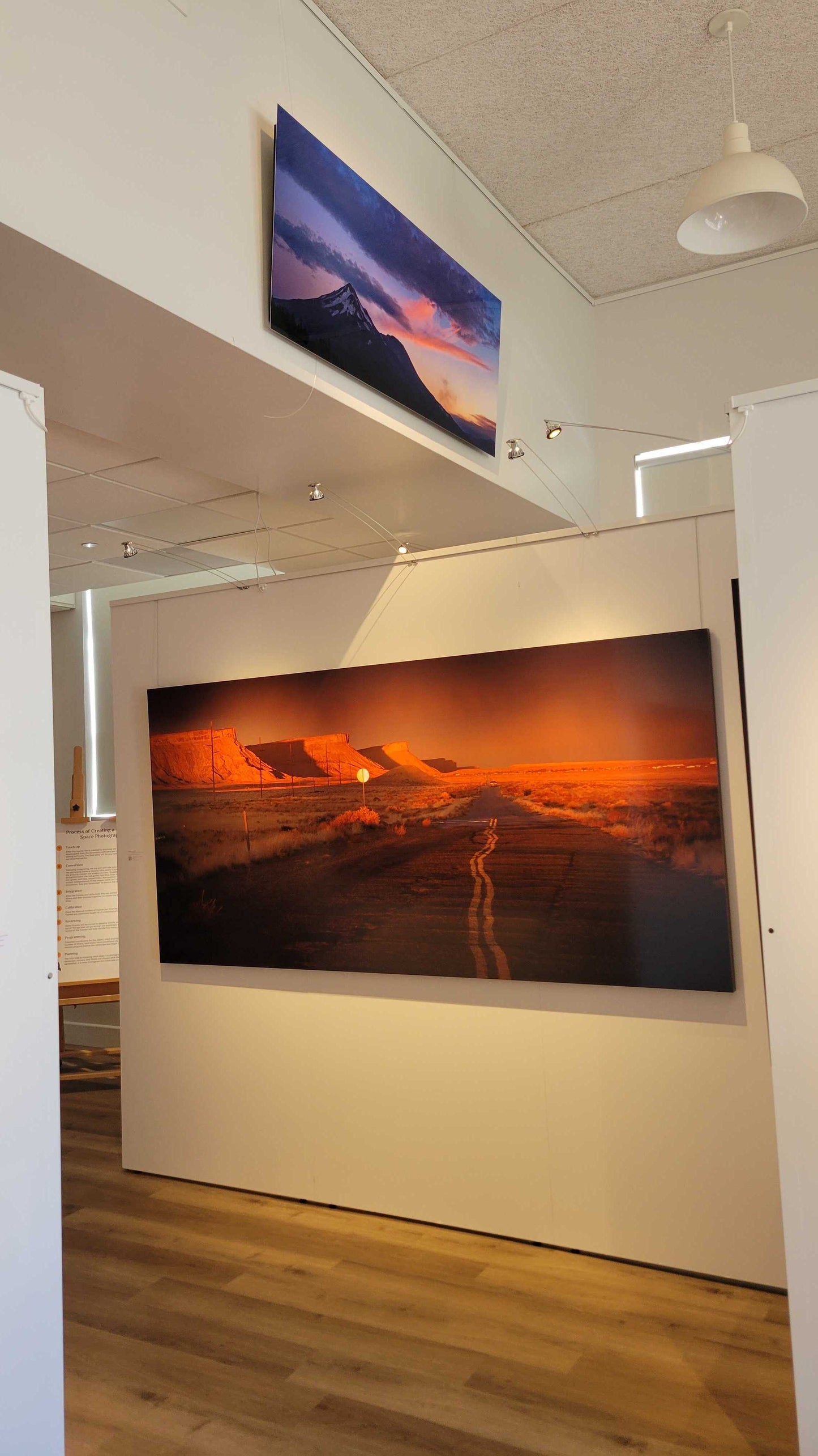 A photograph of the interior view of Slice of Light Gallery showing the two metal prints titled "Mt Shasta, August 2021: Fire Above Smoke" and "Heading Home" on display.