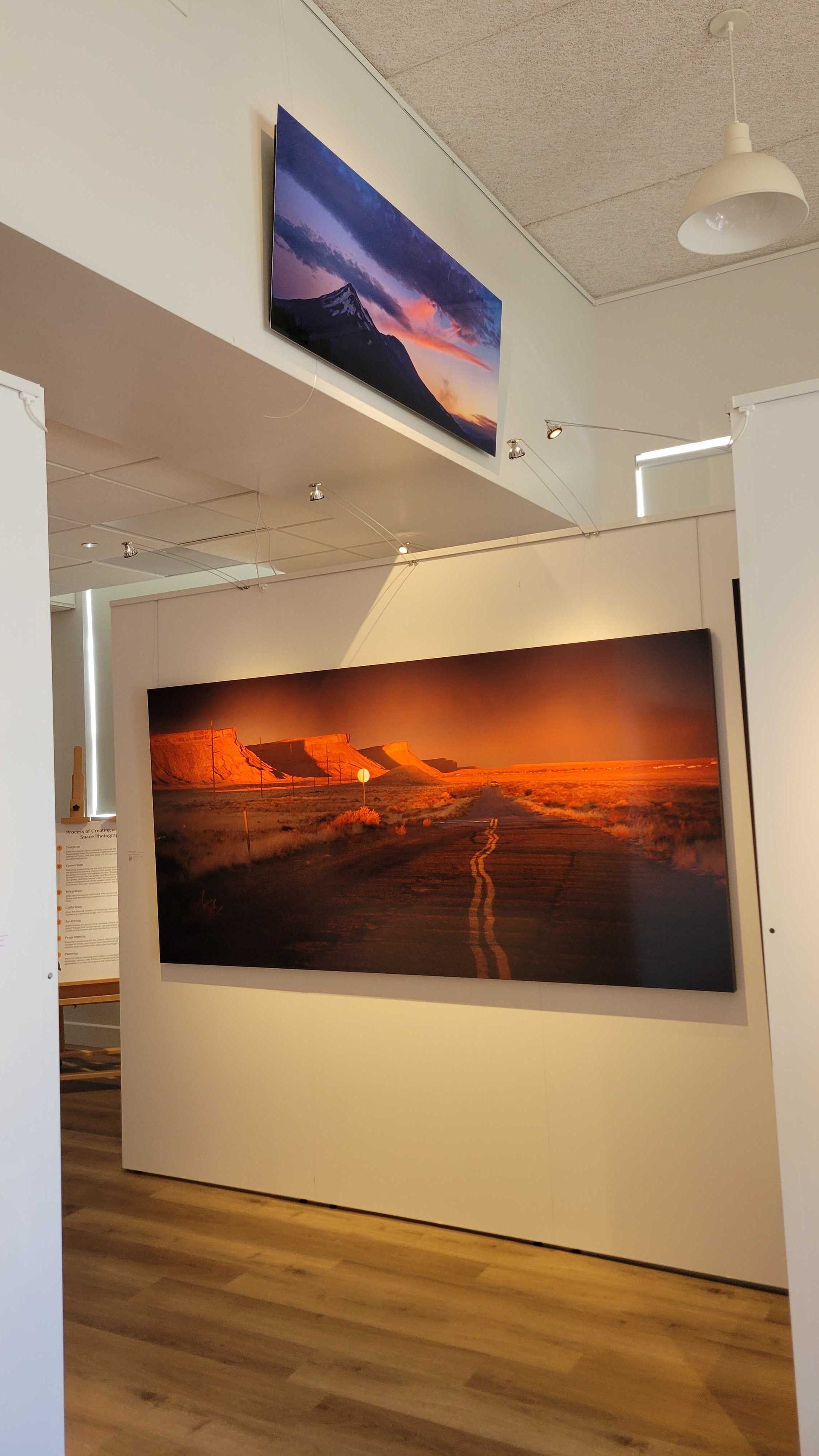 A photograph of the interior view of Slice of Light Gallery showing the two metal prints titled "Mt Shasta, August 2021: Fire Above Smoke" and "Heading Home" on display.