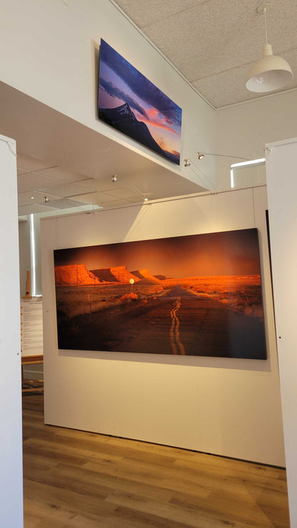 A photograph of the interior view of Slice of Light Gallery showing the two metal prints titled "Mt Shasta, August 2021: Fire Above Smoke" and "Heading Home" on display.